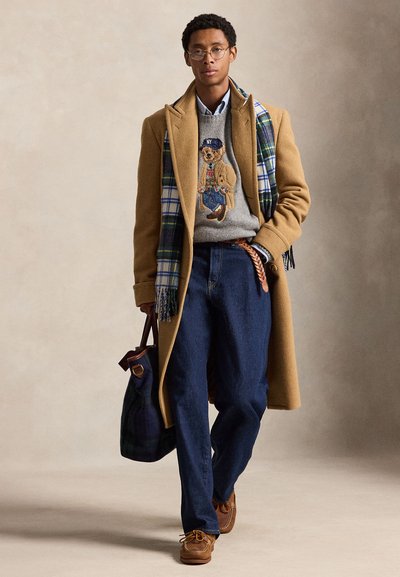 Homme portant un manteau camel, une écharpe à carreaux, un pull gris avec un motif d’ours, un jean bleu, des chaussures marron, portant un sac foncé à motifs, marchant sur un fond uni.
