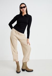 Camisa polo negra de manga larga, pantalones anchos beige y botines de ante marrón con un grueso tacón negro, combinados con gafas de sol negras.
