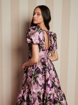 Vestido floral em roxo com flores rosas, mangas bufantes, costas abertas com detalhe de laço e barra em camadas com franjas. Textura de cetim suave.