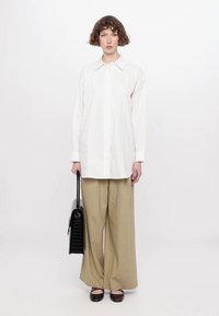 Camicia bianca con bottoni e maniche lunghe, abbinata a pantaloni beige larghi. Gli accessori includono una borsa nera testurizzata e scarpe basse marroni.