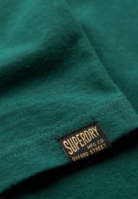 Grøn bomuldsstof, der viser en søm og et sort mærke med guldtekst, der angiver "SUPERDRY MFG CO OXFORD STREET." Glat tekstur.