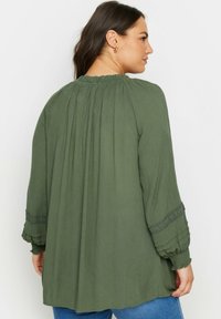 Olivgrön blus med pösiga, samlade ärmar, texturerad tyg och spetsdetaljer vid ärmsluten. Avslappnad passform, bakvy visas.