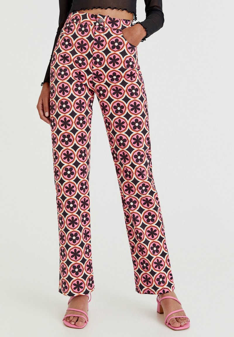 Femme portant un pantalon à taille haute et jambes droites avec un motif floral géométrique rose, noir et orange, associé à des sandales à talons roses.