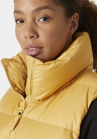 Žlutá péřová vesta s vysokým límcem a zipovým zapínáním. Tkanina je hladká, s prošívanou texturou a lesklým povrchem.