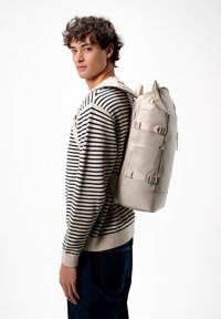 Béžový vodotěsný batoh s rolovacím uzávěrem, nastavitelnými popruhy a přezkami, který má na sobě osoba v pruhovaném svetru.