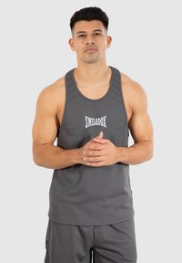 Grijze geribde tanktop met een ronde halslijn, voorzien van de merknaam "SMILODOX" in het wit. Het model staat met de handen gevouwen.