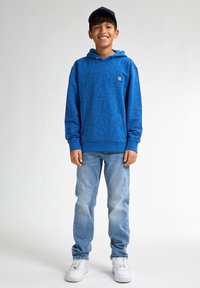 Modrá mikina s kapucí a texturovanými vzory loga, kontrastní šňůrkou, v kombinaci se světle modrými džínami a bílými teniskami. K tomu se dokonale hodí kšiltovka.