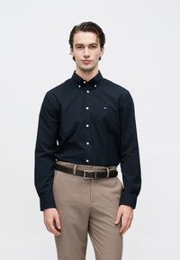 Chemise bleu marine à boutons blancs avec petit logo, associée à un pantalon marron clair et une ceinture sombre. Tissu lisse, coupe ajustée.