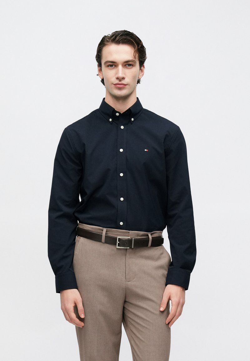 Chemise bleu marine à boutons blancs avec petit logo, associée à un pantalon marron clair et une ceinture sombre. Tissu lisse, coupe ajustée.