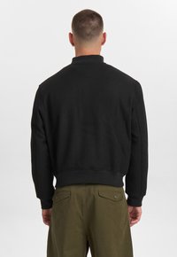 Felpa nera con colletto alto, polsini e vita a costine, caratterizzata da una texture liscia. Mostrata di schiena, abbinata a pantaloni verde oliva.