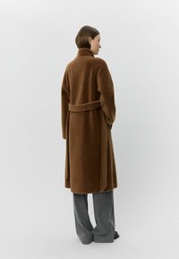 Brauner, langer Mantel aus weichem Stoff mit hohem Kragen und Gürtel, von hinten gesehen. Kombiniert mit grauen, weit geschnittenen Hosen.