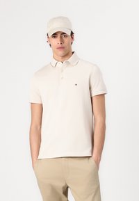 Beige polo shirt med korte ærmer, tre-knaps placket og logo patch på brystet. Kombineret med beige bukser og en lys kasket.
