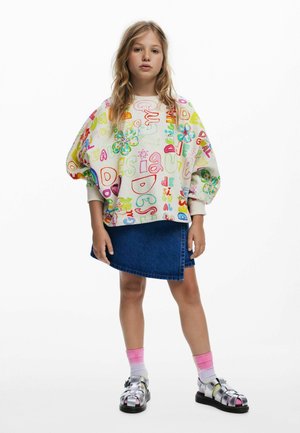 Kleurrijke oversized trui met speelse patronen, spijkerrok en metallic sandalen met roze en witte gradient sokken.