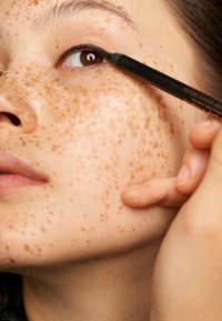 Närbild av ett ansikte med ljus hudton som har framträdande fräknar. En hand applicerar en svart eyelinerpenna längs ögonlocket.