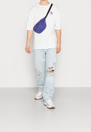 Camiseta blanca oversized, jeans rasgados de color azul claro y zapatillas blancas con suelas de goma. Una pequeña mochila cruzada de color púrpura se lleva sobre el pecho.