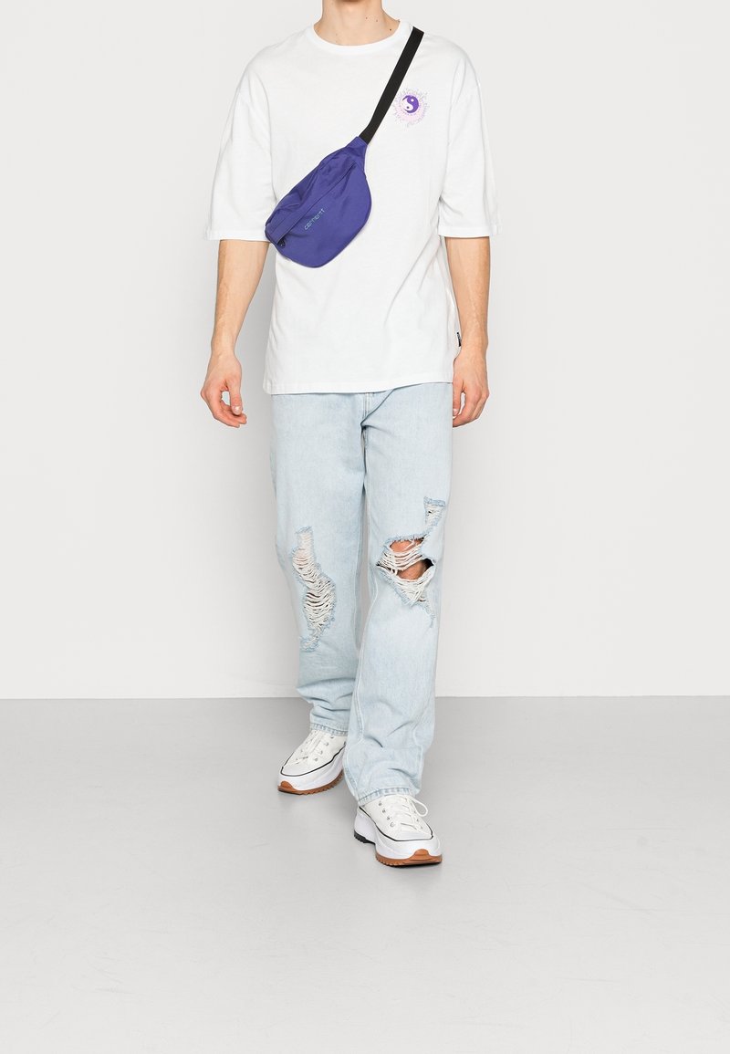 Camiseta blanca oversized, jeans rasgados de color azul claro y zapatillas blancas con suelas de goma. Una pequeña mochila cruzada de color púrpura se lleva sobre el pecho.