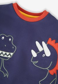 Tricou bleumarin cu desene brodate de dinozauri: un T. rex cu dinți ascuțiți și un triceratops cu colți albi, accente roșii și galbene.