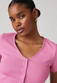 Haut rose à manches courtes en côtes avec un décolleté en V et six petits boutons sur le devant. Texture lisse avec un design ajusté.