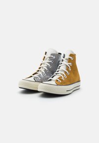 Tenisky s vysokým střihem a bílou podrážkou a tkaničkami, které mají jednu stranu z hnědého kordu hořčičné barvy a druhou stranu se černobílými pruhy.