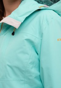 Veste turquoise clair avec une fermeture éclair à l'avant, un col doux et une doublure intérieure texturée. Elle présente des accessoires ajustables et un logo discret.