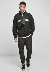 Svart hettegenser med "STARTER"-logo, kombinert med sorte cargo-bukser og sorte joggesko. Inkluderer en liten svart crossover-veske.