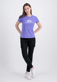 Lilla kortærmet t-shirt med hvidt logo, parret med sorte cargo-bukser og hvide sneakers. Glat stof, tætsiddende design.