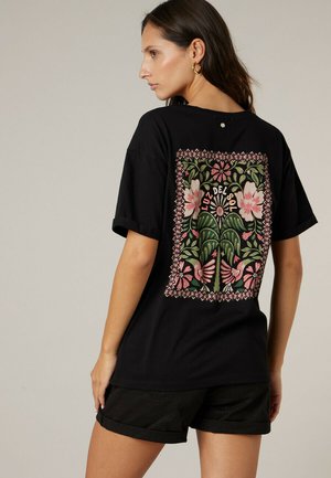Vrouw draagt een zwart T-shirt met een roze en groene bloemenprint en de tekst "Luz Del Sol" op de achterkant, gecombineerd met een zwarte short.