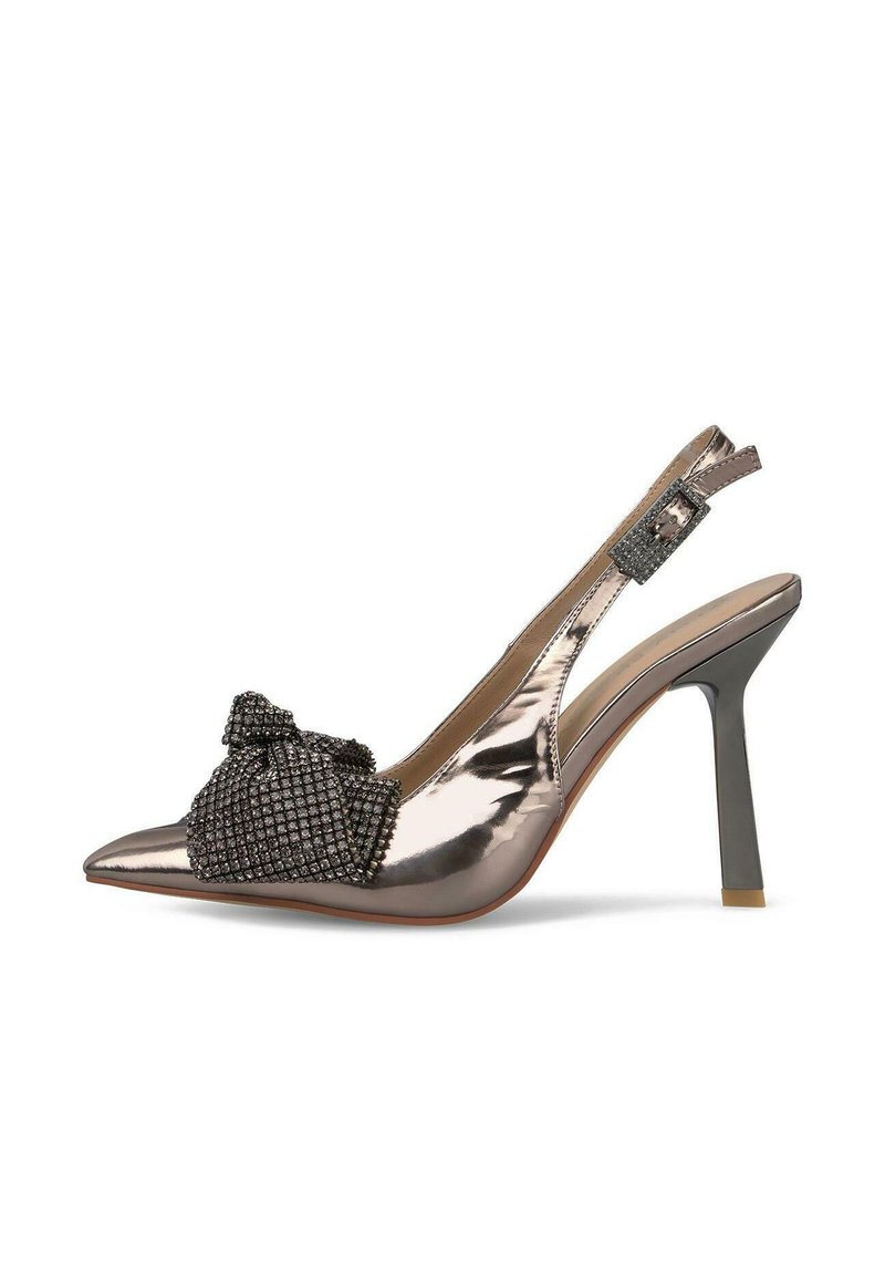 Tacón slingback de bronce metálico con puntera puntiaguda, decorado con un lazo negro texturizado y detalles de tachuelas, que presenta un fino tacón de aguja.