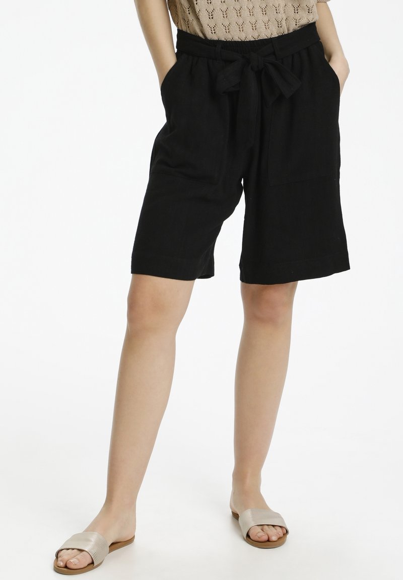 Femme portant un short noir sous le genou avec des poches avant et une ceinture à nouer, un haut en tricot beige et des sandales argentées à glisser, se tenant avec les mains dans les poches.