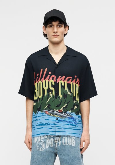 Νέος άνδρας που φοράει μαύρο καπέλο, μαύρο πουκάμισο με κουμπιά με το λογότυπο «Billionaire Boys Club» και γραφικό με βάρκα, καθώς και ανοιχτό μπλε τζιν με παρόμοιο κείμενο.