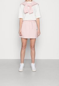 Jupe en coton rose à cordon de serrage associée à un t-shirt blanc surdimensionné. Un sweatshirt rose clair est noué autour du cou. Des baskets blanches aux pieds.