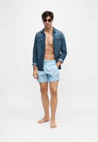 Un homme se tient pieds nus, porte des lunettes de soleil, une chemise en jean ouverte et un short de bain bleu clair sur fond blanc uni.