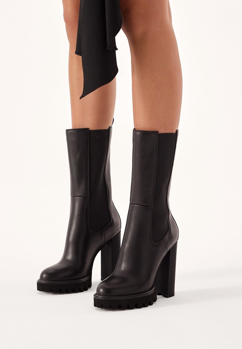 Bottes noires en cuir à la cheville avec un talon bloc épais, des panneaux latéraux élastiques et une semelle en caoutchouc texturée pour une meilleure adhérence. Finition lisse, design élégant.