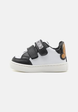 Sneaker bianco e nero per bambino con due cinghie in velcro, testo "MOSCHINO" sulla cinghia superiore e faccia di orsetto color beige sul tallone.