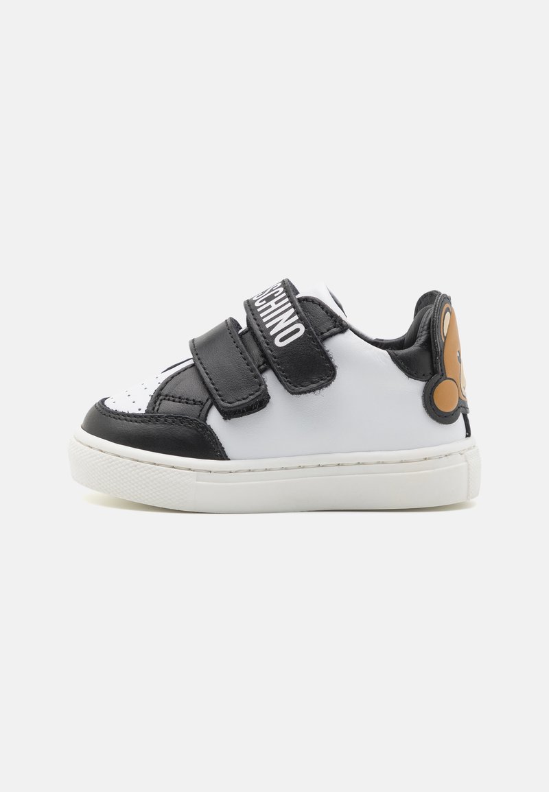 Sneaker bianco e nero per bambino con due cinghie in velcro, testo "MOSCHINO" sulla cinghia superiore e faccia di orsetto color beige sul tallone.