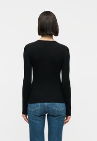 Γυναίκα με ίσια σκούρα μαλλιά, φορώντας εφαρμοστό μαύρο μακρυμάνικο τοπ και μπλε τζιν, που στέκεται με την πλάτη προς την κάμερα.