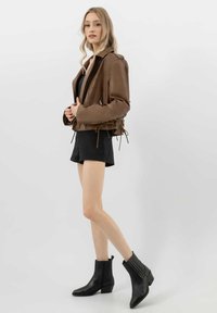 Giacca cropped in pelle marrone con zip laterali e dettagli con lacci, abbinata a una t-shirt nera e pantaloni corti, con stivaletti neri alla caviglia.