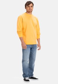 Sudadera amarilla con cuello redondo y puños de canalé, combinada con jeans de mezclilla azul claro y zapatillas de skate negras en una pose de pie.