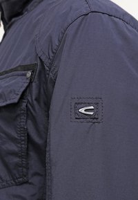 Marineblauwe jas met een ritszak, textuurstof en een zilveren logo-patch op de mouw. Gestikte details langs de naden.