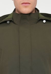 Olive zelená bunda s vysokým golierom, zapínaním na snap gombíky a hladkým povrchom. Dve predné vrecká sú viditeľné.