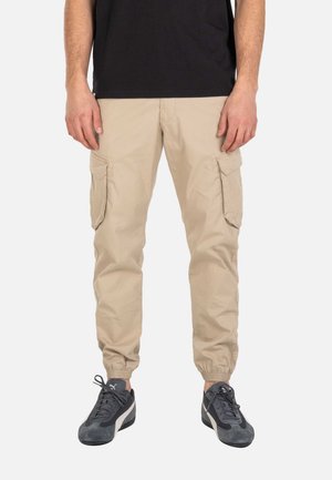 Hombre que lleva pantalones cargo beige con puños elásticos y zapatillas Puma negras y grises, de pie frente a un fondo liso.