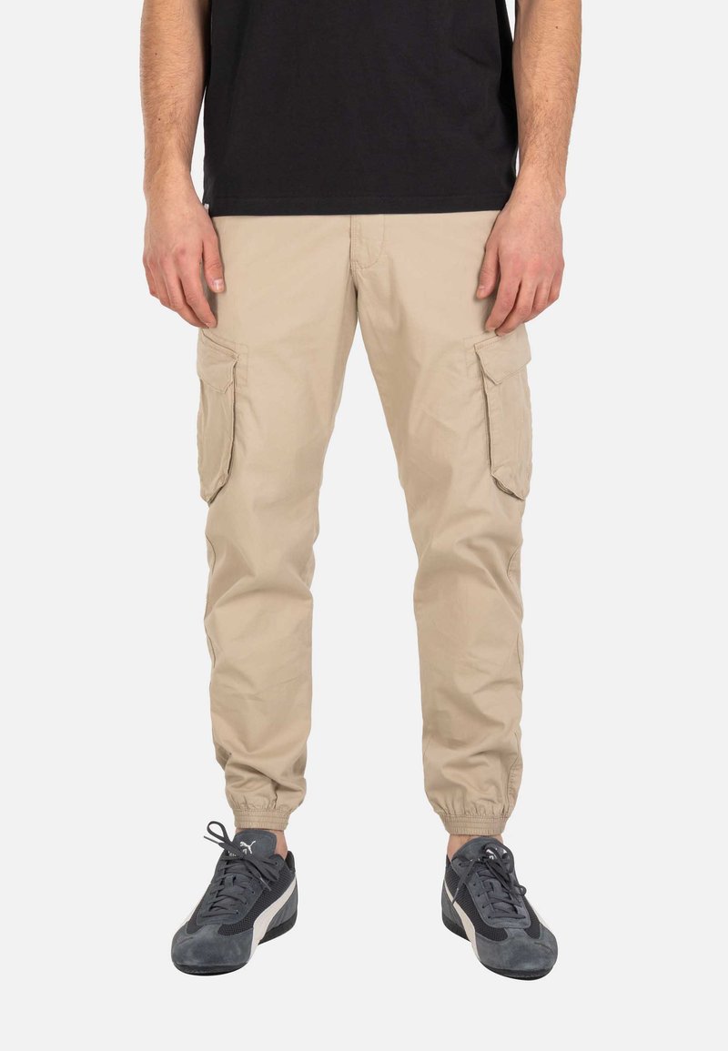 Hombre que lleva pantalones cargo beige con puños elásticos y zapatillas Puma negras y grises, de pie frente a un fondo liso.
