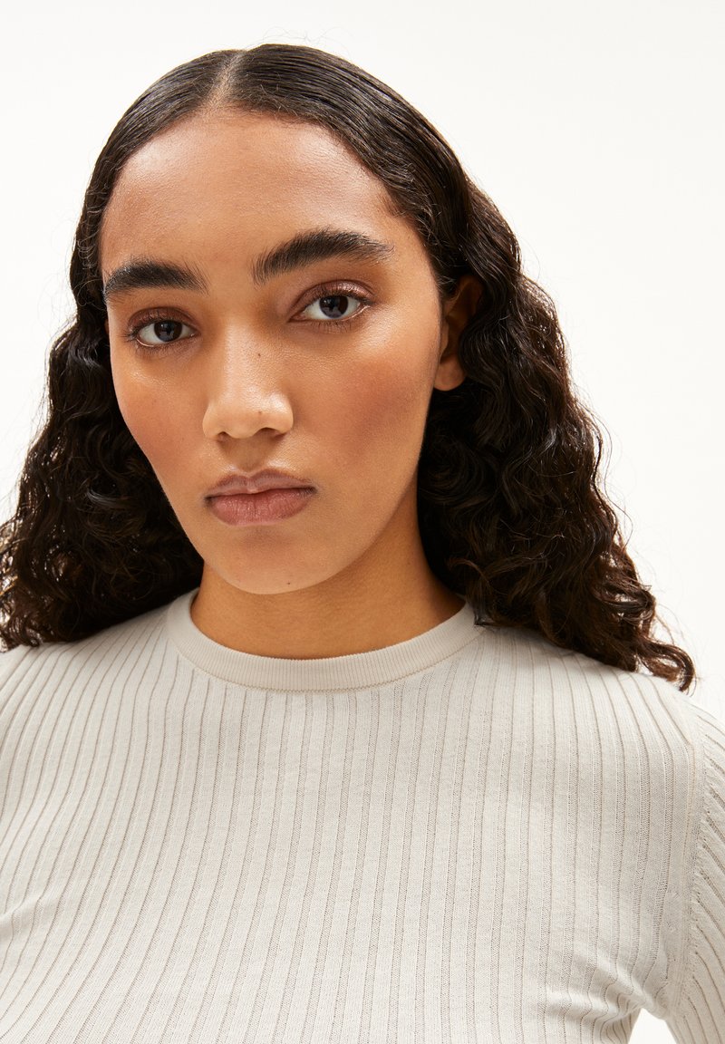 Hellgrau geripptes Oberteil mit Rundhalsausschnitt. Der Stoff hat eine glatte Textur und zeichnet sich durch ein körpernahes Design aus, das den Kragen und die Ärmel betont.