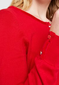 Röd tröja med rund halsringning, ribbade ärmslut och dekorativa knappar på axlarna. Mjuk tygtextur.