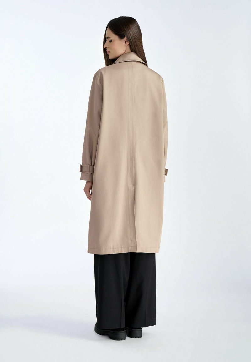 Beige Trenchcoat mit Rückenschlitz, langen Ärmeln und Schnallenakzenten. Über weit geschnittenen schwarzen Hosen getragen, die eine glatte Stofftextur aufweisen.