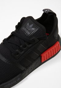 Svart atletisk sko med texturerad stickad överdel, svarta snören och röd gummidetalj på sulan. Har Adidas-logotyp på tungan och sidan.