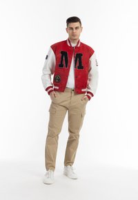 Giacca varsity rossa e bianca con scritta nera "M", dettagli con toppe, pantaloni cargo khaki e sneakers bianche. Design semplice e atletico.
