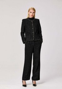 Costume en tweed noir avec une veste ajustée ornée d'accents dorés et de boutons, associé à un pantalon droit et des escarpins en cuir verni noirs.