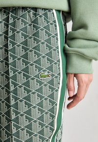 Mão repousada ao lado de calças verdes com padrão, com faixas laterais brancas e verdes e um pequeno logótipo de crocodilo bordado.