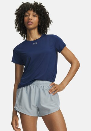 Vrouw poseert met hand op de heup, gekleed in een marineblauw sport-T-shirt en lichtgrijze hardloopshorts tegen een effen witte achtergrond.